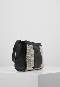 Borsa a tracolla in tessuto intrecciato bianco e nero con dettagli e tracolla in pelle nera, esposta su un piedistallo bianco su uno sfondo grigio chiaro.