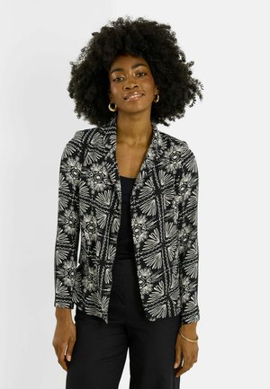 Femme avec coiffure afro portant un blazer à motif floral noir et blanc, un top noir, un pantalon noir, des boucles d'oreilles anneaux dorées et un bracelet, debout.