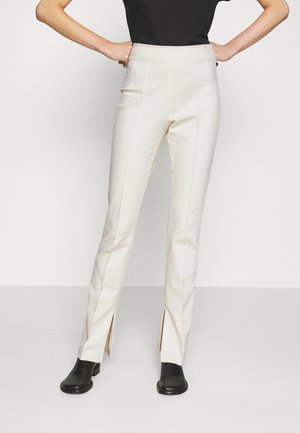 Woman wearing high-waist white pants with front seams and ankle zippers paired with black boots and a black top, hands on hips.