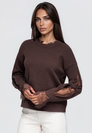 Une femme porte un pull marron foncé orné de dentelle à l’encolure, aux manches et aux poignets, associé à un pantalon blanc sur un fond uni.