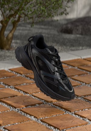 Black athletic shoes featuring a mesh upper, textured detailing, and a thick rubber sole, positioned above a tile and gravel surface.