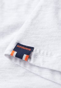 Camiseta de algodón blanca con una etiqueta en azul marino y naranja que presenta "Superdry" en texto negrita. Textura suave con sutiles detalles de costura.
