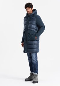 Marineblauer Puffer-Mantel mit Kapuze, der eine Mischung aus glänzenden und matten Texturen, horizontale Nähte und zwei Vordertaschen aufweist. Getragen mit Jeans und Stiefeln.