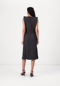 Vestido midi sem mangas, cinza escuro, com ombros estruturados, cintura cingida por um cinto e detalhe de bolso nas costas. Combinado com sandálias de salto pretas.