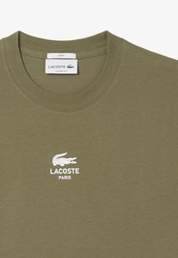 T-shirt in cotone verde oliva con scollatura rotonda, caratterizzato da un logo coccodrillo bianco di Lacoste e la scritta "PARIS" sotto di esso. Design di vestibilità classica.