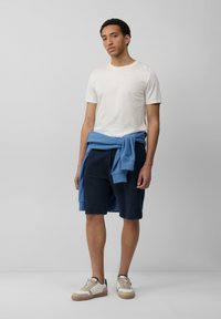 Witte katoenen t-shirt, lichtblauwe sweatshirt om de taille gebonden, marineblauwe shorts en beige sneakers met witte accenten; casual outfit.