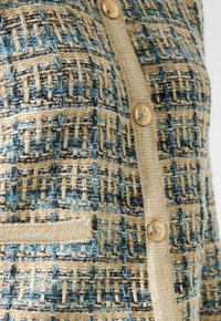 Tejido de punto texturizado con hilos en tonos beige, azul y negro, que presenta un ribete beige y botones decorativos dorados con diseños en relieve.