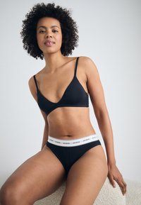 Fekete fehérnemű szett, ami egy háromszög alakú bralette-ből és egy hozzá illő hipster alsónadrágból áll, fehér logózott derékkal. Puha anyag és minimális design jellemzi.
