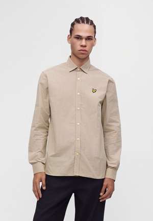 Ung mand med flettet hår iført en beige ternet skjorte med knapper og en gul sommerfugl-logo på brystet samt sorte bukser.
