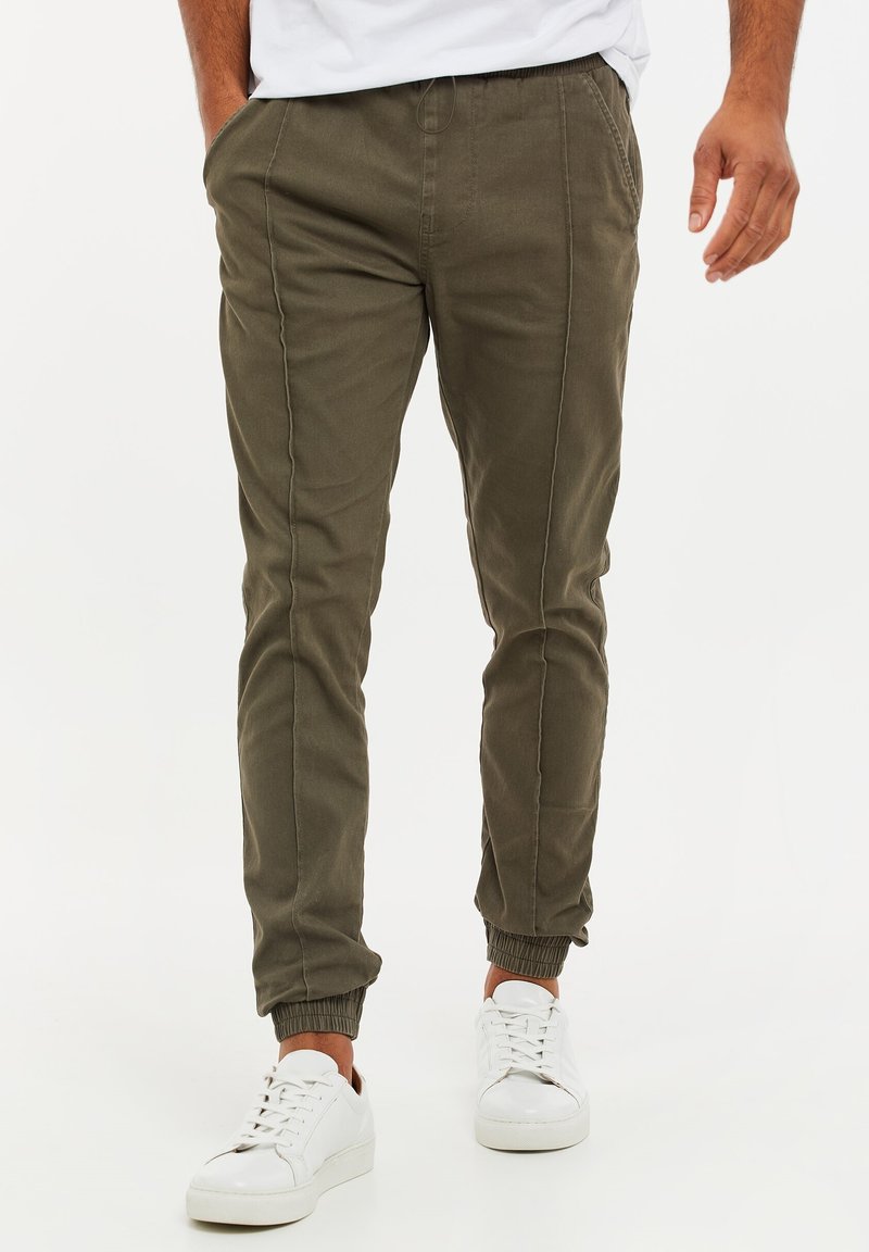 Homme portant un pantalon de jogging vert olive avec des poches latérales et des baskets blanches, marchant vers l'avant sur un fond uni.