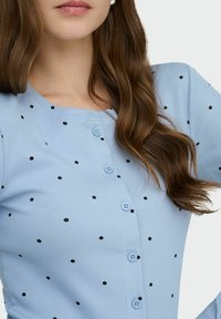 Haut boutonné bleu clair côtelé à pois noirs, avec un col rond et de petits boutons bleus, conçu pour une tenue décontractée.