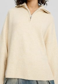 Beige Zip-Up-Pullover mit hohem Kragen, lockerer Passform, geripptem Saum und weicher Textur, ausgestattet mit einem einzelnen Reißverschluss am Kragen.