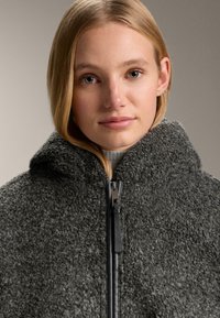 Temno siva jopica iz flisa s teksturirano površino, ki ima visok ovratnik in črno zadrgo. Material deluje mehak in topel.