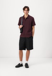 Korte mouw bordeaux shirt met een gestructureerd ruitpatroon, gecombineerd met zwarte shorts en zwarte schoenen. Model draagt een kleine zwarte tas.
