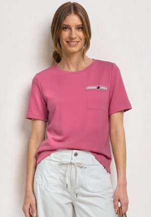 Ung kvinde iført en pink kortærmet skjorte med en dekoreret lomme og hvide snorbukser, stående og smilende.