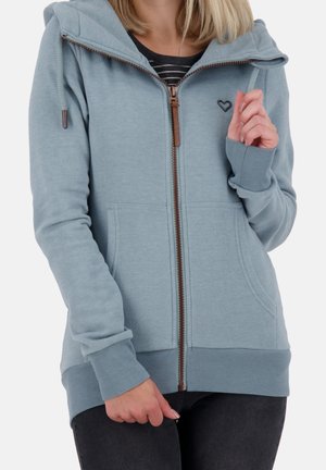 Femme portant un sweat à capuche zippé bleu clair avec un petit logo cœur près de la poitrine, associé à un pantalon foncé et une chemise rayée en dessous.