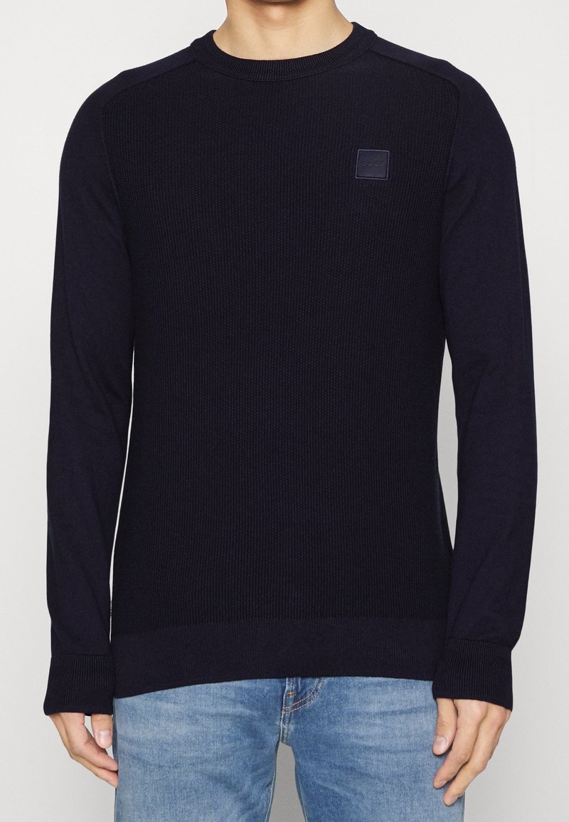 Homme portant un pull en maille côtelée bleu marine uni à col rond avec un petit patch logo carré sur la poitrine gauche, associé à un jean bleu clair.