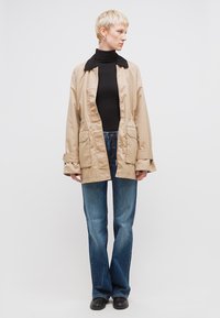 Άτομο με κοντά ξανθά μαλλιά που φοράει μπεζ μπουφάν πάνω από μαύρο ζιβάγκο, μπλε τζιν και μαύρα παπούτσια, στέκεται μπροστά σε απλό φόντο.