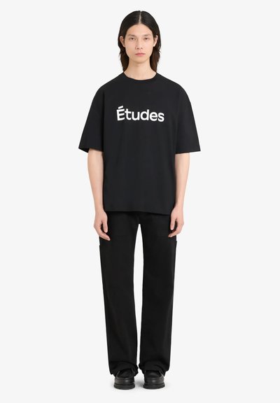 T-shirt en coton noir à manches courtes avec un grand texte blanc "Études". Associé à un pantalon noir à jambes larges et des chaussures noires.