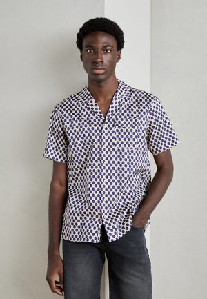 Camisa de manga corta en blanco con patrón de puntos azules. Presenta un cuello tipo polo, cierre de botones en la parte delantera y un bolsillo. Se lleva con jeans oscuros.