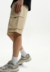 Pantaloni scurți cargo bej cu două buzunare mari pe laterale, confecționați din țesătură de bumbac. Purtați cu șosete albe și teniși în nuanțe neutre.