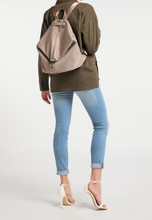 Beige rugzak van glad materiaal, voorzien van een rits, verstelbare banden en een driehoekige vorm. Draagt over een groene jas en spijkerbroek.