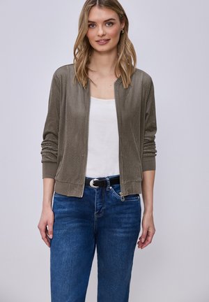 Joven mujer con chaqueta gris perforada con cremallera sobre una camiseta blanca y vaqueros azules, posando frente a un fondo claro.