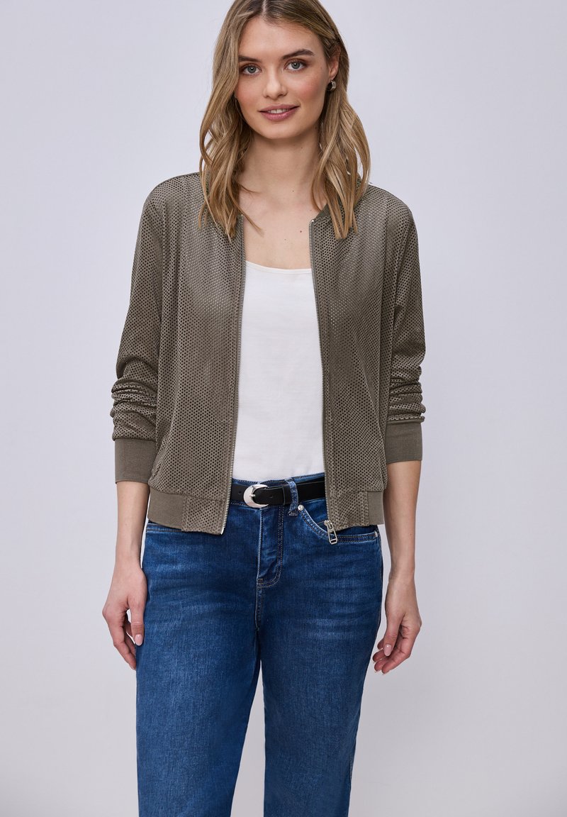 Ung kvinde iført grå perforeret lynlåstrøje over hvid top og blå jeans, stående mod en lys baggrund.