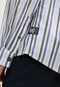 Overhemd met knopen, voorzien van verticale strepen in wit, blauw en zwart. Katoenen stof met een geweven textuur. Merktag aan de onderzijde.