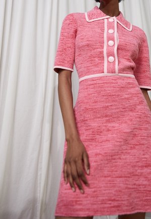 Kobieta w dopasowanej różowej sukience z krótkimi rękawami, białym wykończeniem, ostrym kołnierzem i czterema dużymi guzikami na przednim panelu.