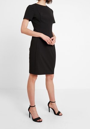 Robe noire ajustée à manches courtes, encolure structurée et coutures verticales. Associée à des sandales à talons hauts noires avec lanières.
