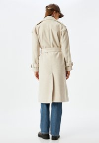 Abrigo trench de lana beige con cintura ceñida, hombreras y correas en los puños. Cuenta con una abertura en la parte posterior. Combinado con unos vaqueros de pierna ancha azules.