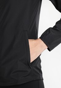 Černá lehká bunda s texturovanou tkaninou, úzkým střihem a zipovou postranní kapsou. Dlouhé rukávy s elastickými manžetami pro pohodlí.