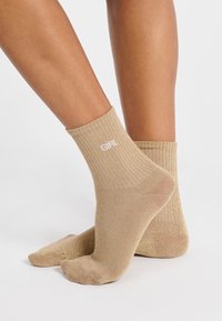 Calcetines de tobillo de hilo beige con un brillo metálico y el texto blanco "GIRL" en el puño. Textura suave con un ajuste ceñido alrededor del tobillo.