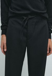 Pantaloni da jogger neri con vita regolabile, tasche frontali e una texture liscia, indossati con una maglietta a maniche lunghe nera abbinata.