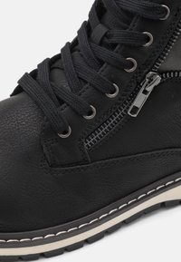 Botín de cuero negro hasta el tobillo con acabado texturizado, que presenta cordones negros, cremalleras plateadas y una suela blanca cosida con un patrón de banda de rodadura robusto.