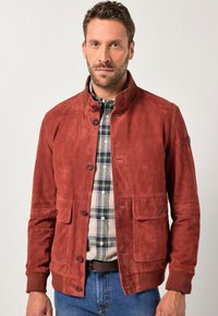 Chaqueta de ante roja con cuello, cierre de botones, dos bolsillos delanteros y puños acanalados. Llevada sobre una camisa de cuadros.
