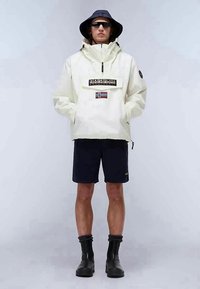 Anorak leve, em creme, com bolso com fecho, com um patch da bandeira norueguesa, usado sobre calções escuros e botas pretas até ao tornozelo, combinado com um chapéu de balde.