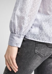 Blusa viola chiaro con motivo floreale, tessuto strutturato e maniche arricciate, dotata di due bottoni sul polsino, abbinata a jeans grigi.