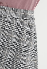 Pantalones de cuadros ajustados hechos de una tela texturizada. Con una cintura alta que presenta patrones en negro, blanco y gris, complementados por un sutil borde beige.
