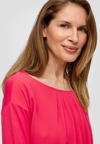 Rosa Bluse mit weitem Ausschnitt, die vertikale Falten und fließenden Stoff aufweist. Das Model hat lange Haare und trägt dezente Ohrringe.