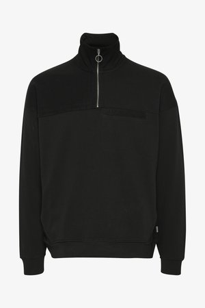 Sweat-shirt noir à zip quart avec un panneau supérieur texturé, associé à un pantalon cargo vert olive avec des poches latérales et des baskets noires.