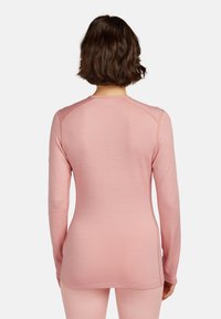 Kobieta z krótkimi brązowymi włosami nosi dopasowaną różową bluzkę z długim rękawem i pasujące spodnie, ukazana z tyłu na jednolitym tle.