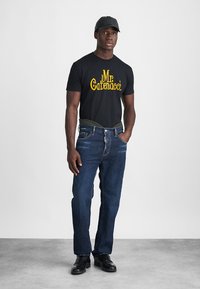 T-shirt noire avec un texte graphique jaune, jeans bleu foncé et chaussures noires. Le modèle porte une casquette noire assortie et se tient les mains dans les poches.