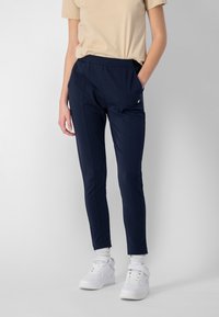 Persona che indossa pantaloni slim-fit blu navy, una maglietta a maniche corte beige, calze bianche e scarpe da ginnastica bianche, in piedi con una mano in tasca.
