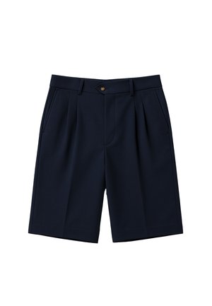 Mężczyzna w czarnych szortach do kolan, białych skarpetkach z czarnym napisem oraz czarno-białych mokasynach, stojący na jednolitym tle.