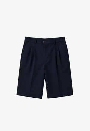 Man draagt zwarte knielange shorts, witte sokken met zwarte tekst en zwart-witte loafers en staat tegen een eenvoudige achtergrond.