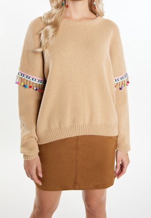 Woman wearing a beige sweater with colorful pom-pom trim on sleeves and a brown suede skirt, standing against a white background.