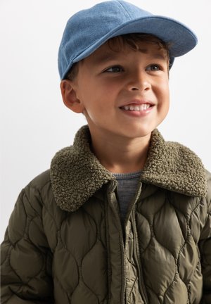 Jeune garçon portant une casquette bleue et une veste matelassée verte avec un col texturé, souriant et regardant sur le côté devant un fond uni.