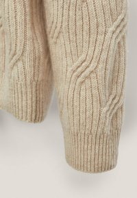 Beiger Strickpullover mit Zopfmuster. Der Stoff zeichnet sich durch eine dicke Rippenstruktur und eine weiche Textur aus, die die Details an Ärmel und Saum hervorhebt.